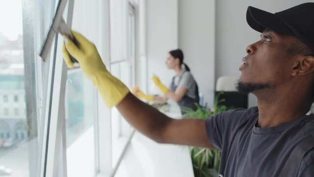 Side view selection focus slowmo of young Black man cleaning window in modern office using detergent spray and spatula