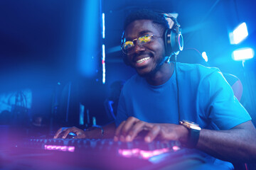 African young male gamer enjoys computer video game, laughs and smiles while playing strategy online © Parilov