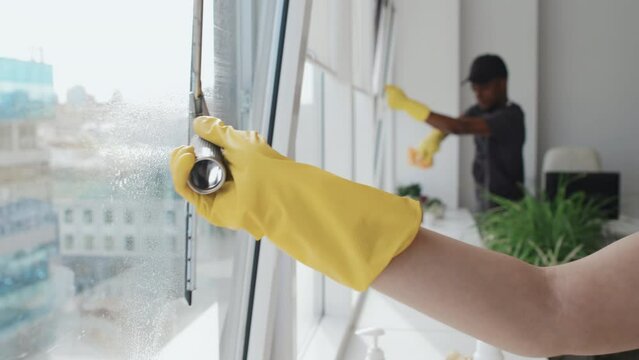 Selective focus slowmo of young Caucasian woman and her Black co-worker providing cleaning service in modern office spraying detergent on windows and wiping it with spatula and cloth