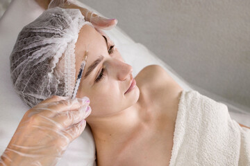 A young woman receives an injection of anti-aging botox filler in the forehead by a professional...