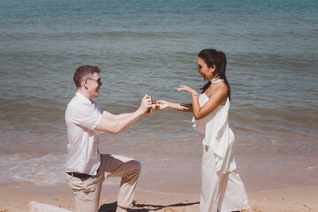 Multicultural love couple concept, Man making proposal with engagement ring and gift box to his woman at the sea beach on valentine day, Caucasian man and Asian women.