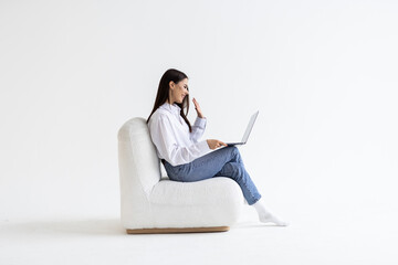 Cheery woman in headphones having online video call on laptop, sitting on chair against white wall,...
