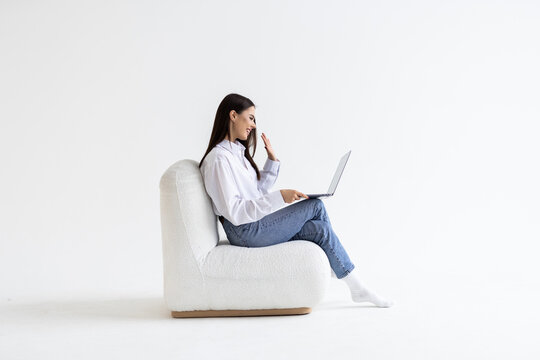 Cheery Woman In Headphones Having Online Video Call On Laptop, Sitting On Chair Against White Wall, Full Length. Millennial Lady Participating In Webinar, Communicating Remotely On Internet