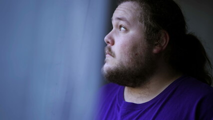 One contemplative overweight man standing by window looking outside by curtain. Pensive expression of a casual chubby guy staring outdoors