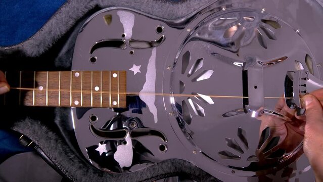 Closeup of a man&rsquo;s hands pulling a new wound string through the bridge and up the neck of a shiny, metal body, resonator acoustic guitar, lying in its case.
