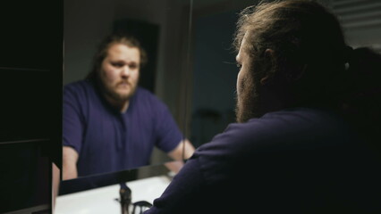 Dramatic person standing in front of bathroom mirror looking at reflection suffering from problems. One pensive serious overweight man facing himself
