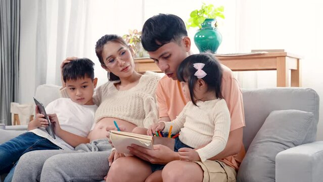 4K, family love warmth. Asian parents are lounging on sofa in living room of house and their son and daughter drawing on coloring book with mom and dad guiding next to woman. Pregnancy, big belly.