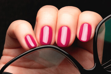 Female hand with beautiful manicure - viva magenta, pink nails with glasses on black background closeup