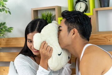 Young happy lover married Asian couple teasing with pillow on white bed. Eye looking husband and...