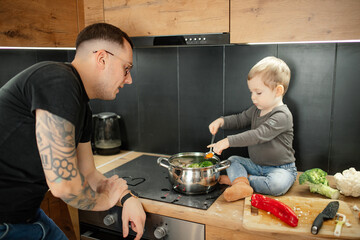 Mature man in eyewear with tattoo on arm teach little son to cook in kitchen. Help father cook vegetable organic soup