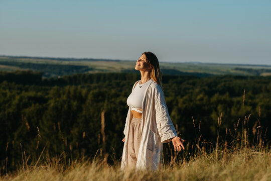 Happy Young Woman Meditating and Catching Sunlight Outdoors at Sunset with Scenic Landscape on the Background
