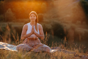 Happy Young Woman with Folded Hands and Closed Eyes Sitting on the Mat and Meditating Outdoors at Sunset