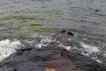 Naklejka premium Lots of small waves hitting red and black beach rocks in Eira aka Eiranranta, Helsinki, Finland. Sunny summer day by the Baltic Sea. Color image showing the power of nature, the sea, and clean Nordic 