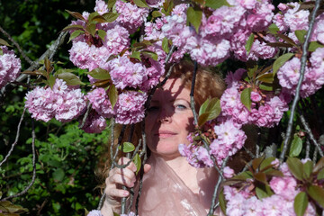 sensual portrait of a sexy, smiling mature, best age redhead woman, hidden behind pink flower tree blossoms in spring awakening april, sakura