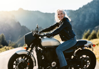 Fototapeta na wymiar smiling middle-aged woman white haired driving a motorcycle on a mountain road. Generative AI