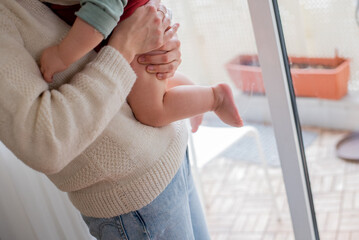 Person holding a baby. Mother holds the child in her arms. Woman is standing near the window. Child with bare legs.