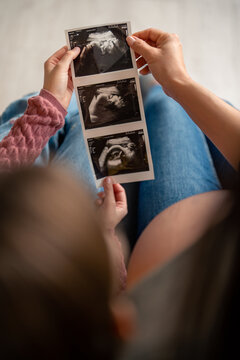 Series of ultrasound baby pictures in hands of unidentifiable pregnant mother and her little daughter. Mother and daughter holding and looking together at sonogram images of future family member.