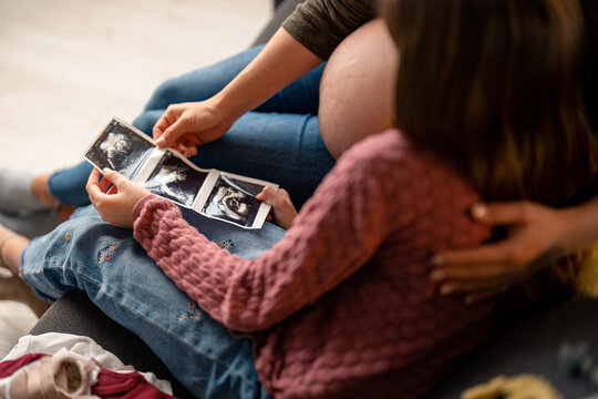 Ultrasound baby photo in hands of pregnant mother and little girl. Mother and daughter sitting together, holding sonogram image of unborn baby.