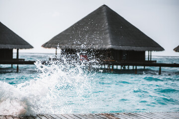 The shot is taken with a selective focus on the water splashing against a wooden footpath, leading to an overwater bungalows with an elegant design featuring thatched roofs, in the Maldives