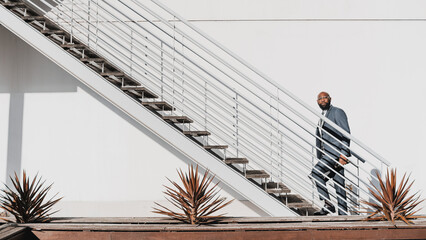 An African businessman starting to climb up the white staircase in front of the white wall, the man is wearing a grey suit in a minimalist aesthetic set with cactus decorating the scene