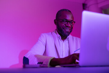 a black man in a white suit and glasses works at a laptop. a man in glasses works in the office in the evening