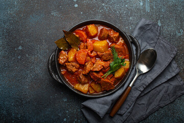 Beef meat stew with potatoes, carrot and delicious gravy in black casserole pot with bay leaves and fresh green herbs with spoon on black dark rustic concrete background from above .