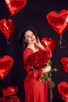 Portrait of a brunette woman with a large bouquet of red roses on a background of heart-shaped balloons.