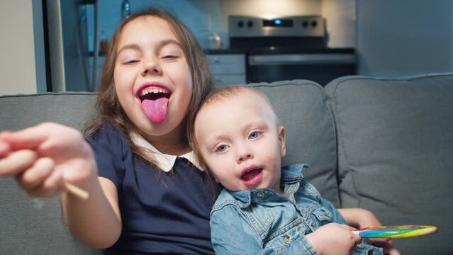 Positive little girl and baby boy licking a lollipop, enjoying a sugar treat and looking at the camera while sitting together on a couch against the background of the kitchen. - Powered by Adobe