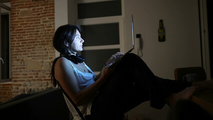 Woman working at night at home in front of laptop computer in the dark. Computer screen glowing on person face while browsing internet online © Marco