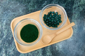 spirulina powder and tablets in glass bowls on a wooden tray with a wooden spoon on a green marble background.