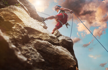 Muscular climber man in protective helmet abseiling from cliff rock wall using rope Belay device and climbing harness on evening sunset sky background. Active extreme sports time spending concept.