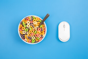Colored breakfast cereal in a bowl and computer mouse on a blue background, breakfast and work concept, conceptual minimalism, flat lay.