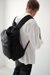 The guy with a bagpack on his shoulders is poaing  on a white background. The teenager  is wearing a white T-shirt and black jeans - obrazy, fototapety, plakaty