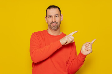 Smiling happy man 40s in casual red pullover pointing index fingers to the side in mock-up workspace area isolated on yellow colored background studio portrait. People lifestyle concept