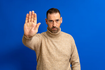 Hispanic handsome man with beard wearing a beige turtleneck sweater over isolated blue background...