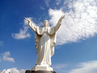 Statua del Sacro Cuore di Gesù su cielo azzurro punteggiato da nuvolette bianche.