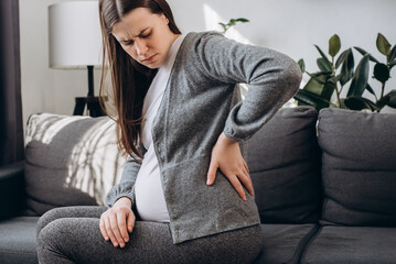 Tired young pregnant woman suffers from back pain sitting on grey sofa in living room at home, future mother suffer from spine muscle ache backache. Concept pregnancy, motherhood, health and lifestyle