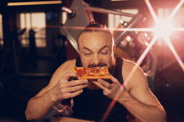 A young man with a beard is happy to eat pizza in the gym