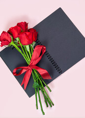 Red rose and blank black paper book on pink background. Copy space.