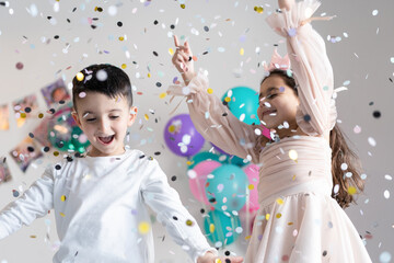 Smiling children with outstretched hands near falling confetti on party background.