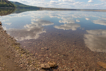 Lake surrounded by a rocky shore, mountains with forest on background.reflection of white clouds in clear water, rocky bottom
