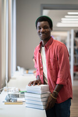 Young cheerful African American man librarian smiling at camera while standing with pile of books...