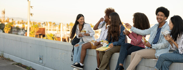 group of multiracial young people eating while sitting outdoors