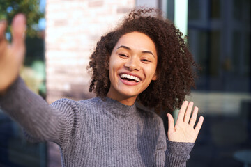 Fototapeta premium Attractive African American woman smiling a toothy smile, waving hello with her hand, expressing positive emotions while looking camera on a smartphone. Video call concept