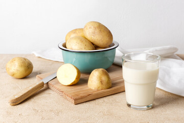 Potato milk, poured into a glass, stands next to a ceramic bowl containing several unpeeled potatoes. Close-up, horizontal orientation.