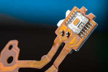 Closeup of optical sensor on electronic printed circuit board and flex ribbon cables on dark blue background. Small orange die in transparent micro chip on PCB of dismantled digital CD/DVD disc drive.