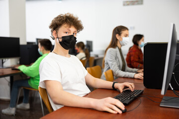 Teenage boy in face mask sitting at table and using computer during lesson.