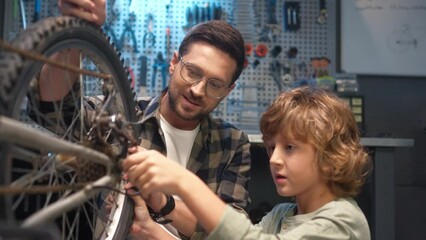 Portrait of young handsome caring caucasian dad and little cute son fixing bike gear at modern garage with tools. Beautiful loving father teaching boy to repair bicycles looking at camera smiling.