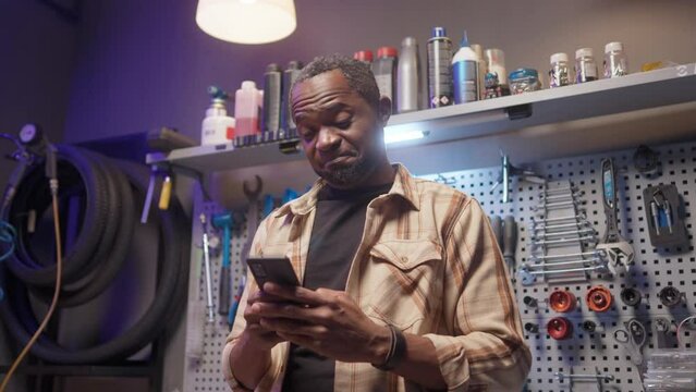 Attractive professional African male repairman having break after fixing bicycles looking at smartphone texting online smiling. Handsome skillful African American mechanic at modern garage with tools.