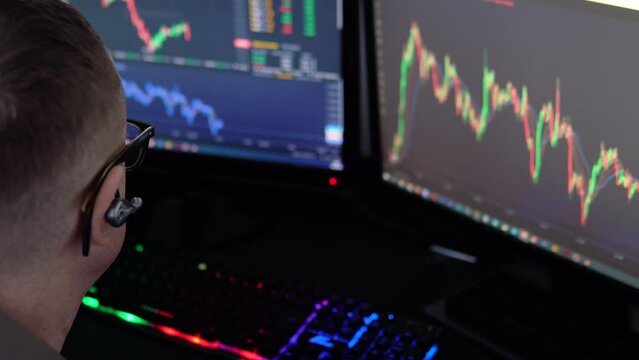 Exchange stock market. Back view of young professional trader in eyeglasses pointing on the data on computer screen with pen and holding Bitcoin in one hand while working his modern office